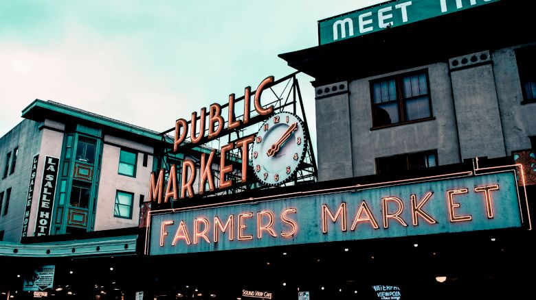 The image shows a neon sign for "Public Market" and "Farmers Market" between two buildings, with part of a sign reading "Meet the" visible on the roof.