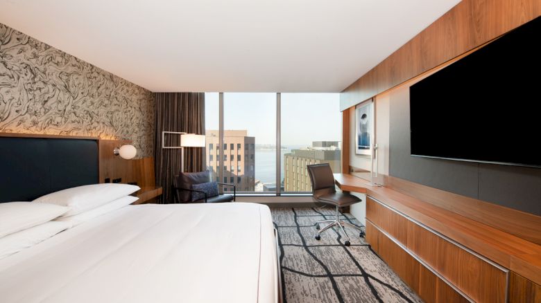 A modern hotel room with a large bed, desk, chair, wall-mounted TV, and a window offering a city view with buildings and water.