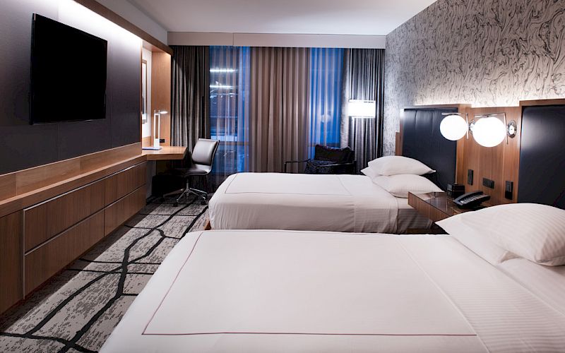 A modern hotel room features two double beds, a large TV, a desk with a chair, and stylish decor, including patterned wallpaper and ambient lighting.