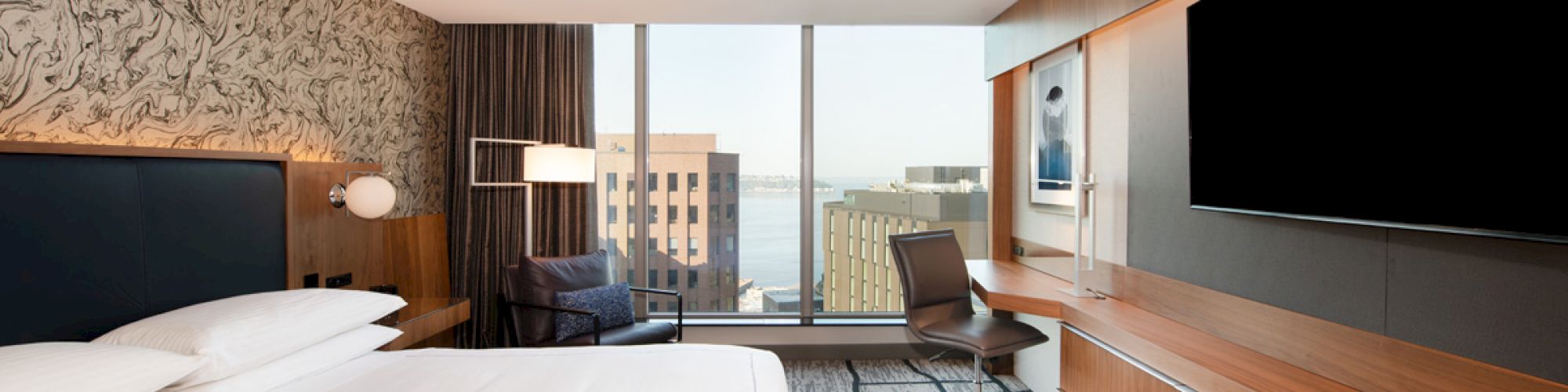 A modern hotel room with a large bed, desk, chair, TV, and a window with a city view and distant waterfront.