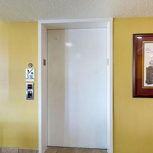 The image shows an elevator door next to a framed picture on a yellow wall. There are buttons and a sign for the elevator on the wall.