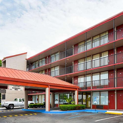 The image shows a mid-sized hotel with a red roof, multiple floors, and balconies. There is a covered entrance with a van parked outside.