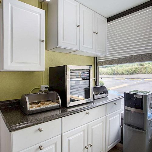 A kitchenette with cabinets, a countertop, a toaster and a microwave oven. There's a water dispenser and a window with blinds in the background.