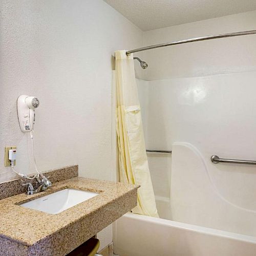 A bathroom with a granite countertop sink, wall-mounted hairdryer, and a walk-in bathtub with a shower curtain and grab bar, no people visible.