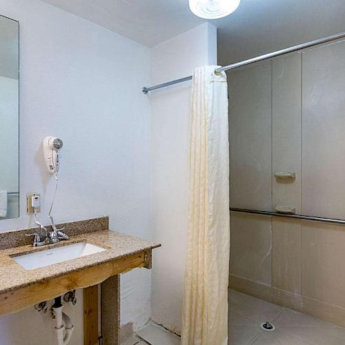 A bathroom with a sink, mirror, hair dryer, and a shower area with a curtain. The sink area appears unfinished or undergoing renovation.