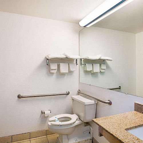 The image shows a clean bathroom with a toilet, sink, countertop, mirror, towel rack with folded towels, and a wall-mounted grab bar.