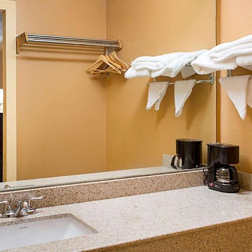 A bathroom counter with a sink, a large mirror, a coffee maker, and neatly folded towels, with a clothes rack and hangers seen in the background.