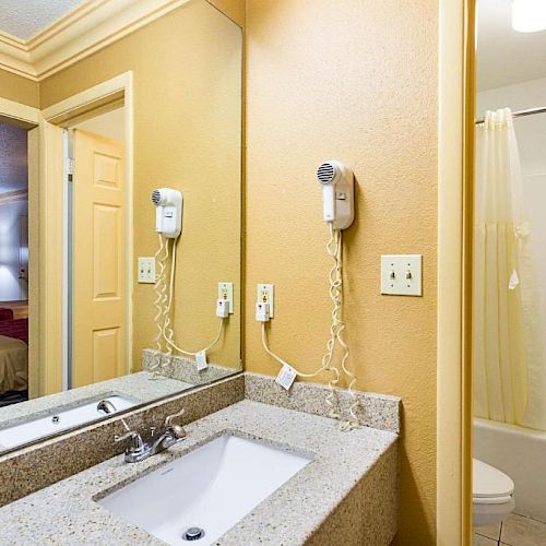 The image shows a bathroom with a granite countertop, sink, mirror, hair dryer, and a shower-tub combination. A bedroom is visible through the doorway.