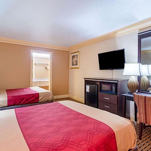 This image shows a hotel room with two beds, a wall-mounted TV, a desk with a mirror, and a chair. The beds have red and beige bedding.