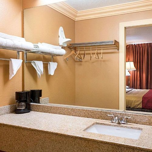 A hotel bathroom vanity with a sink, towels, a hairdryer, and a coffee maker. The reflection in the mirror shows a bedroom with a bed and lamp.