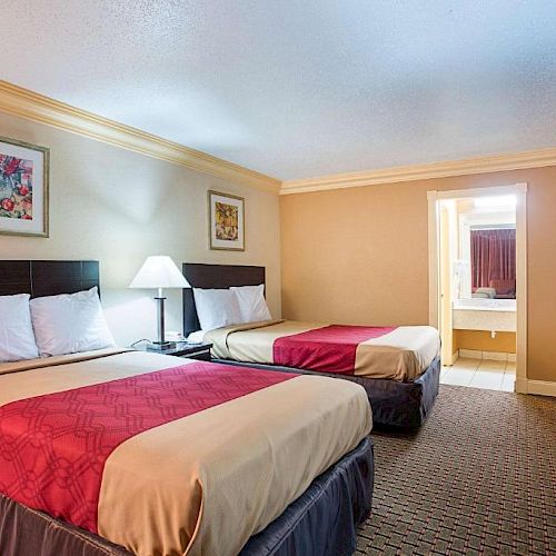 A hotel room with two double beds, a lamp between them, framed artworks on the wall, a TV cabinet, and a bathroom visible in the background.