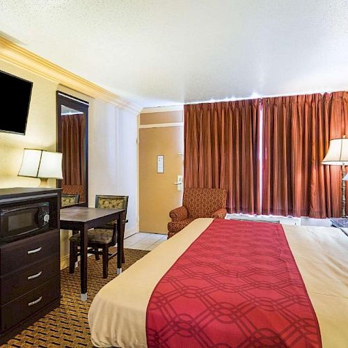 The image shows a hotel room with a bed, a TV, a desk with a chair, and lamps. The room has brown curtains and a patterned carpet.