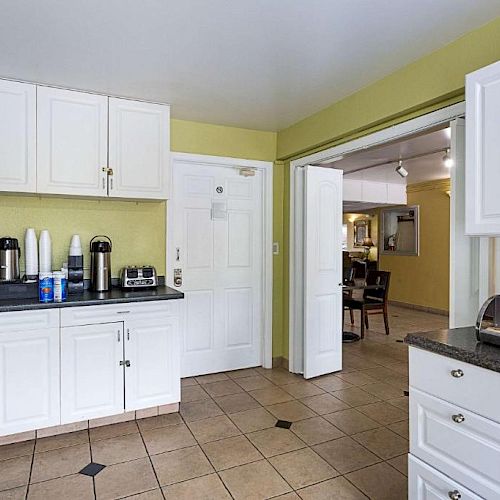 A kitchen with white cabinets, a coffee station, and a pastry display on the countertop; an open doorway leads to a dining area.