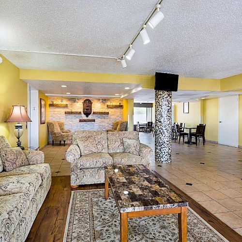 The image shows a spacious living room with yellow walls, patterned sofas, a marble coffee table, and a decorative column, leading to a dining area.