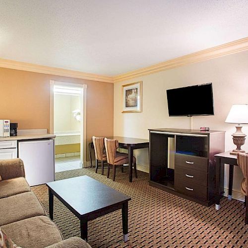 A hotel room features a sofa, a coffee table, a flat-screen TV, a desk, chairs, a mini-fridge, and a microwave with warm lighting.