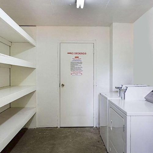 This image shows an empty laundry room with shelves on one side, a door with a sign, and a washer and dryer on the right side.